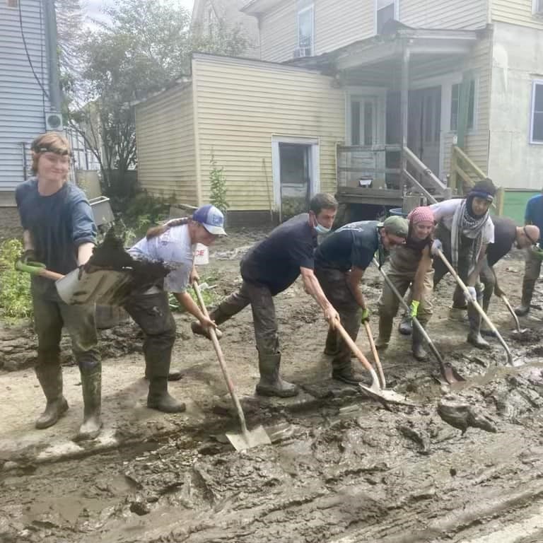 A group of volunteers from Barre Up, holding shovels at the edge of a sidewalk. They all pose and smile at the camera, shoveling mud in unison.