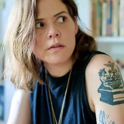 A headshot of Bianca Stone. She is wearing a black tank top and has a tattoo on her arm, and looks off into the distance as if surprised