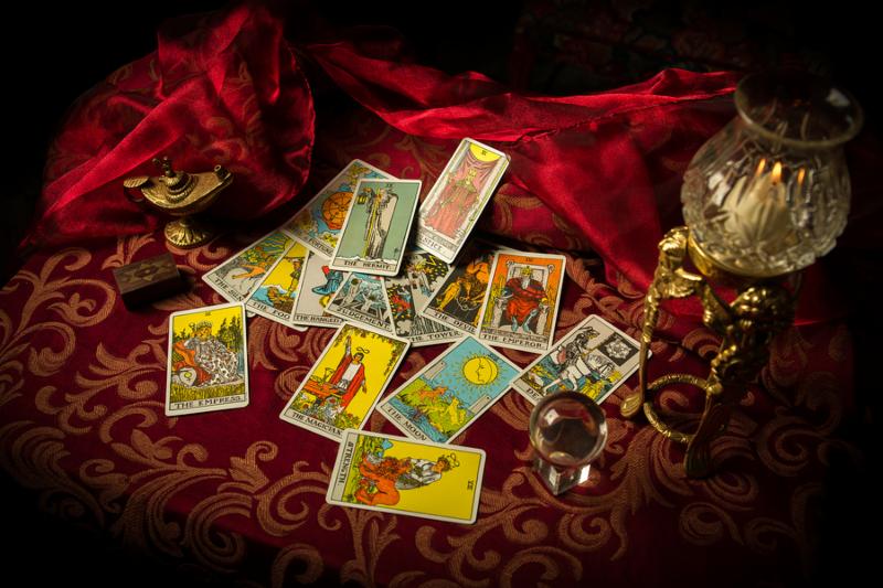 A pile of tarot cards lie scattered and spread across a table top surrounded by multiple occult items.