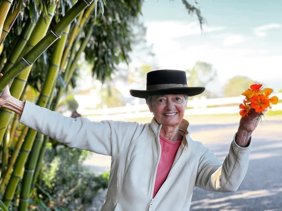 A senior woman wearing a hat and a big smile holds onto a bamboo plant and a bouquet of bright flowers