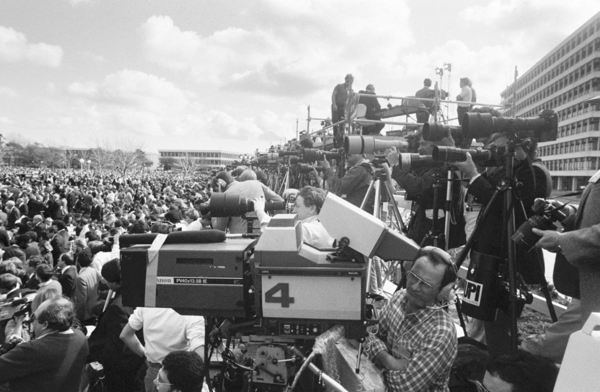 Row upon row of photographrs and videographers line the mall at NASA’s Johnson Space Center where hundreds of attendees have gathered for the STS-51L memorial. The buildings of the Center are seen on the right and in the distance on the left.