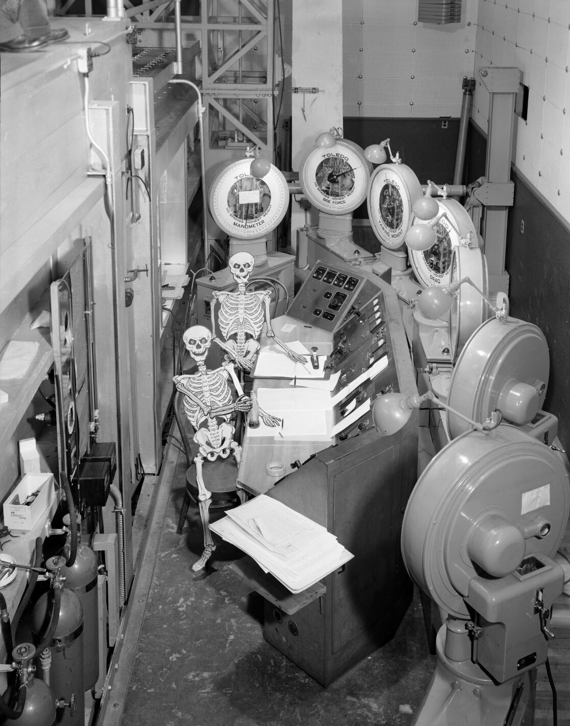 Overhead view of a wind tunnel control room. Two skeletons made of paper sit in the chairs at the console. 