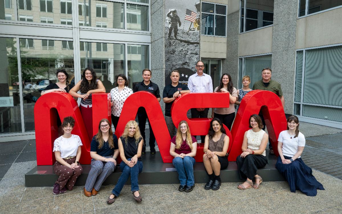 The NASA History Office staff assemble around the NASA worm logo sculpture outside NASA Headquarters