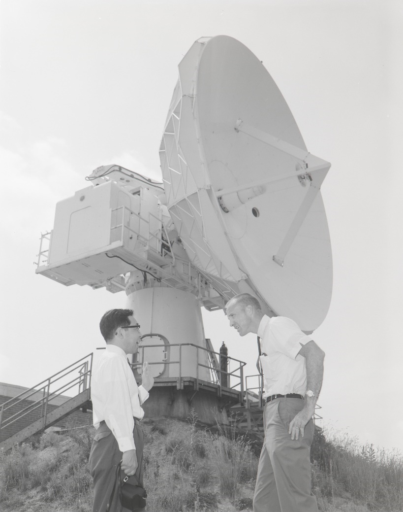 Two men in the foreground face one another as they talk. Behind them on a hill is a large tracking antenna.