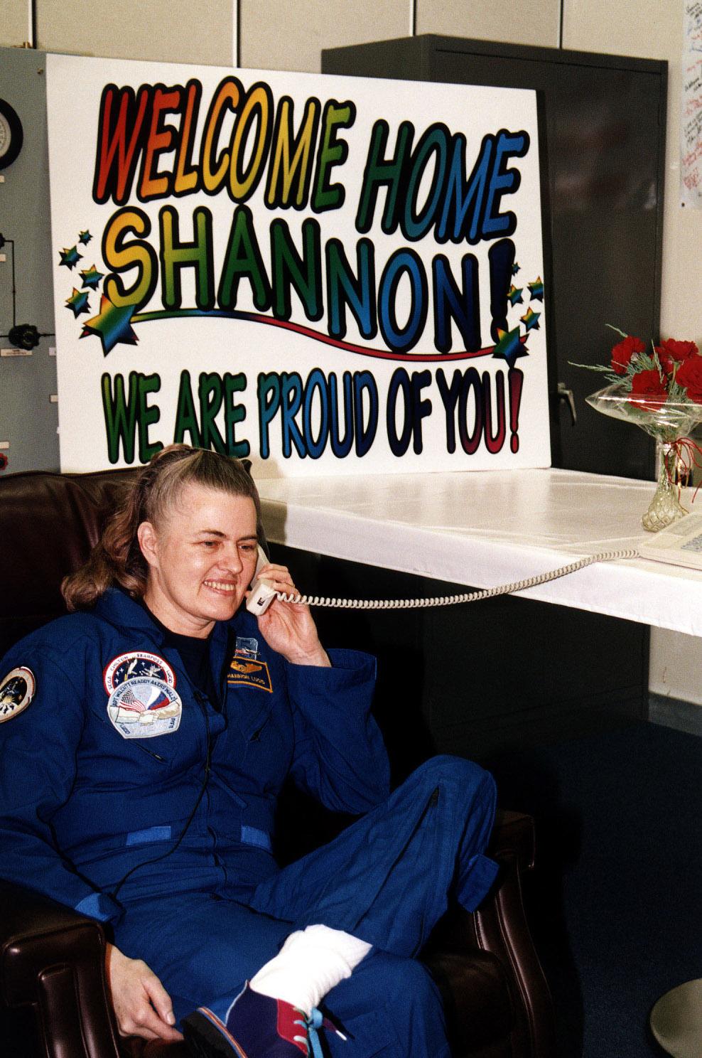 Shannon Lucid talks on the phone seated in front of a banner that reads Welcome Home Shannon! We are proud of you!