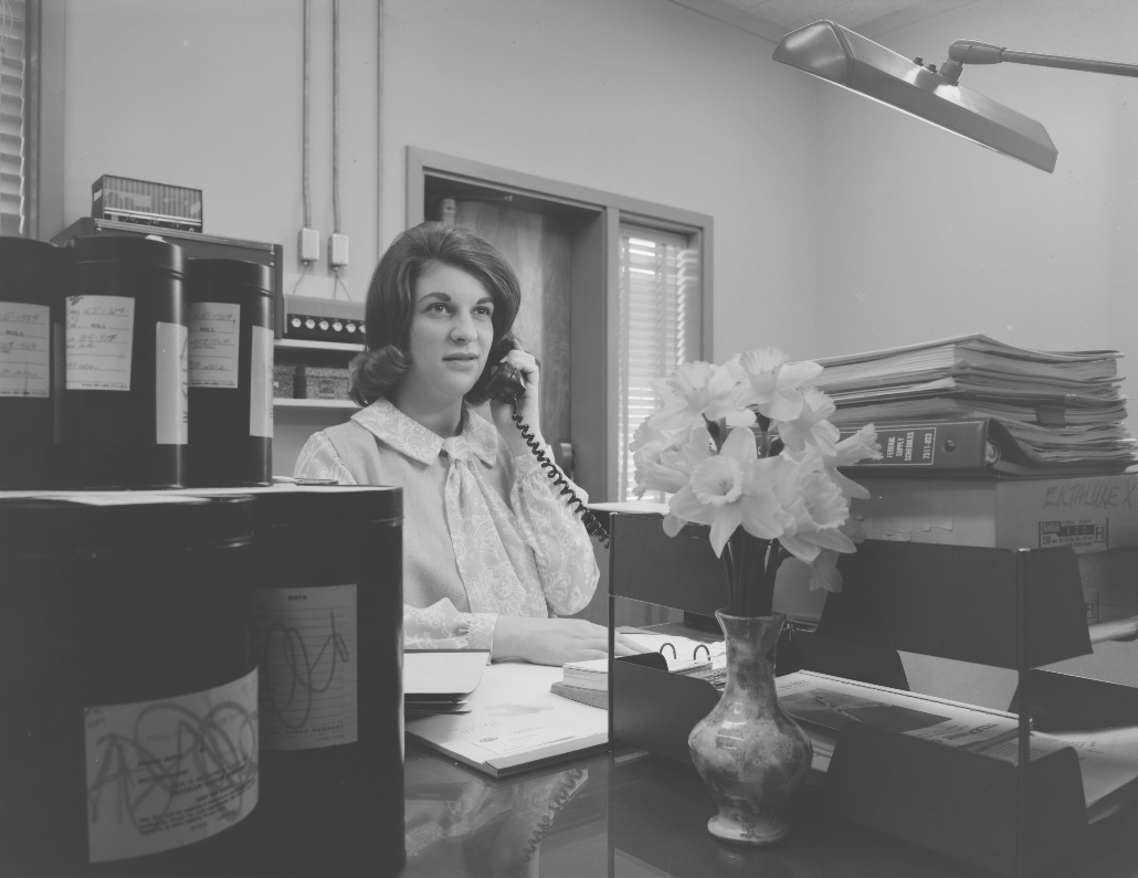 A woman sits at a desk in an office with a phone receiver at her ear.