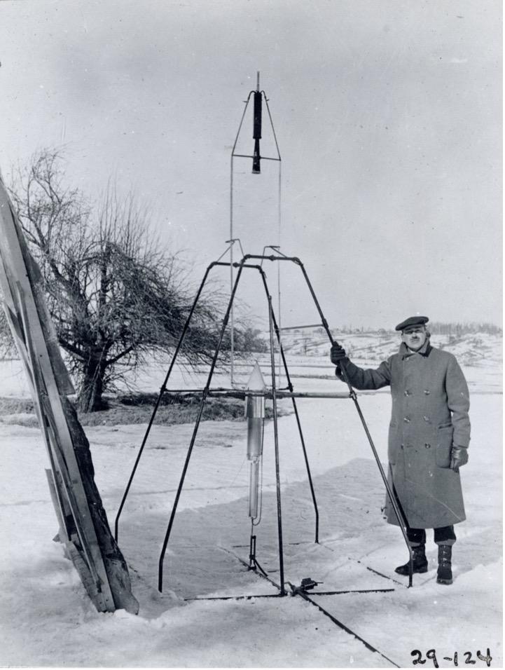 On a snowy March 16, 1926, Dr. Robert H. Goddard rests his hand on the testing frame supporting his liquid fuel rocket at Ward Farm in Auburn, Massachusetts. A wooden door is propped up at an angle next to the frame where Goddard’s assistant, Henry Sachs, later sheltered after lighting the rocket. 