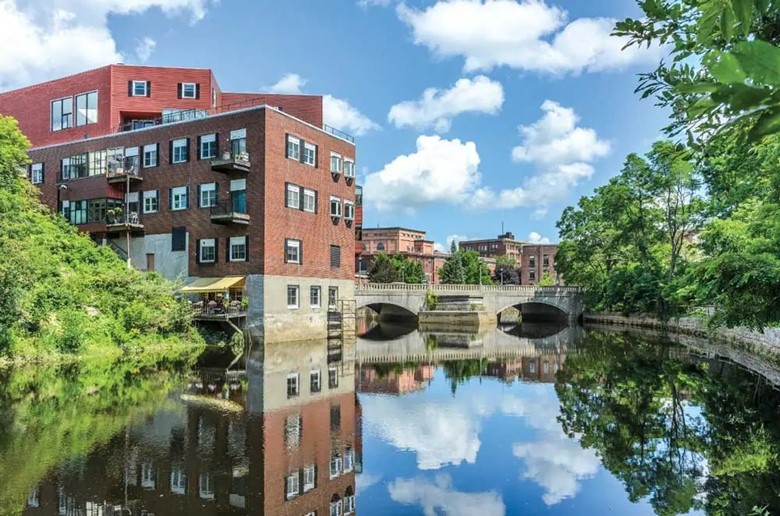 Placid waterway in Greater Bangor on a clear, sunny day reflects clouds in the sky and building in the background