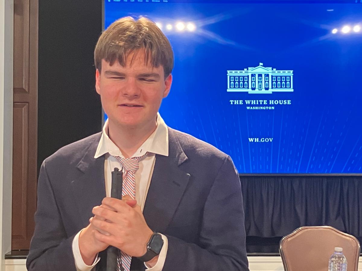 A young man holding a white cane stands in front of a blue screen that reads White House.gov
