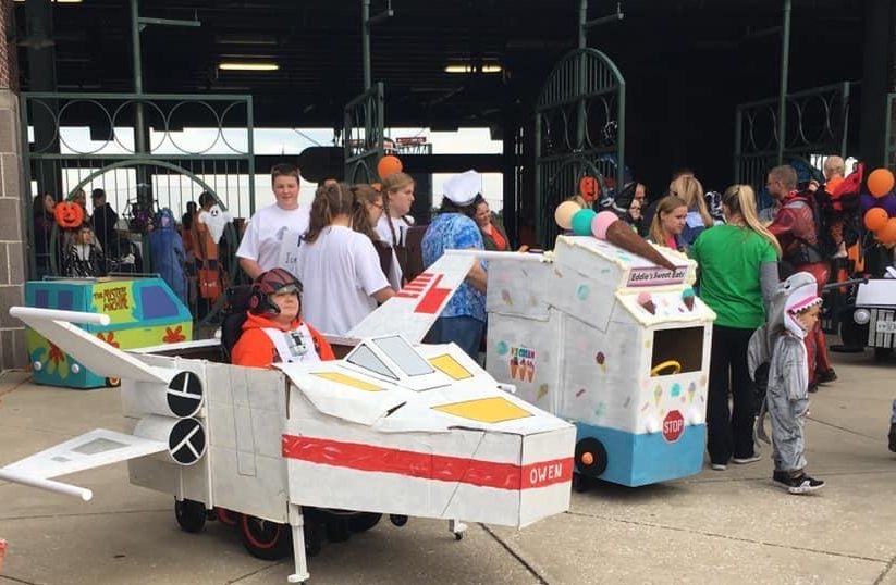 Child has Star Wars X-wing fighter costume around his wheelchair.  Next to him is ice cream truck costume.