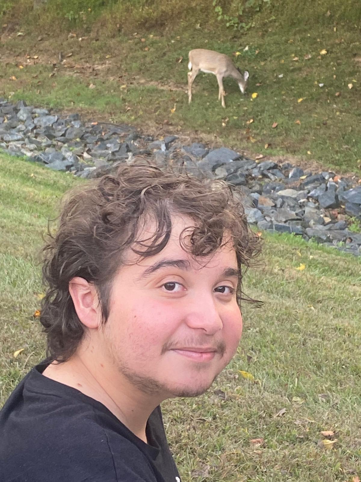 A young man smiles at the camera. A deer is grazing in the field behind him. 
