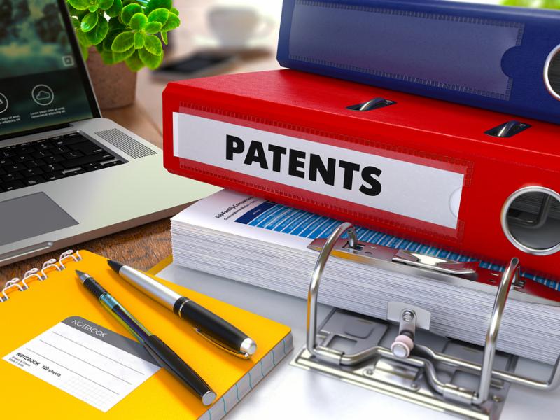Red Ring Binder with Inscription Patents on Background of Working Table with Office Supplies, Laptop, Reports. Toned Illustration. Business Concept on Blurred Background.