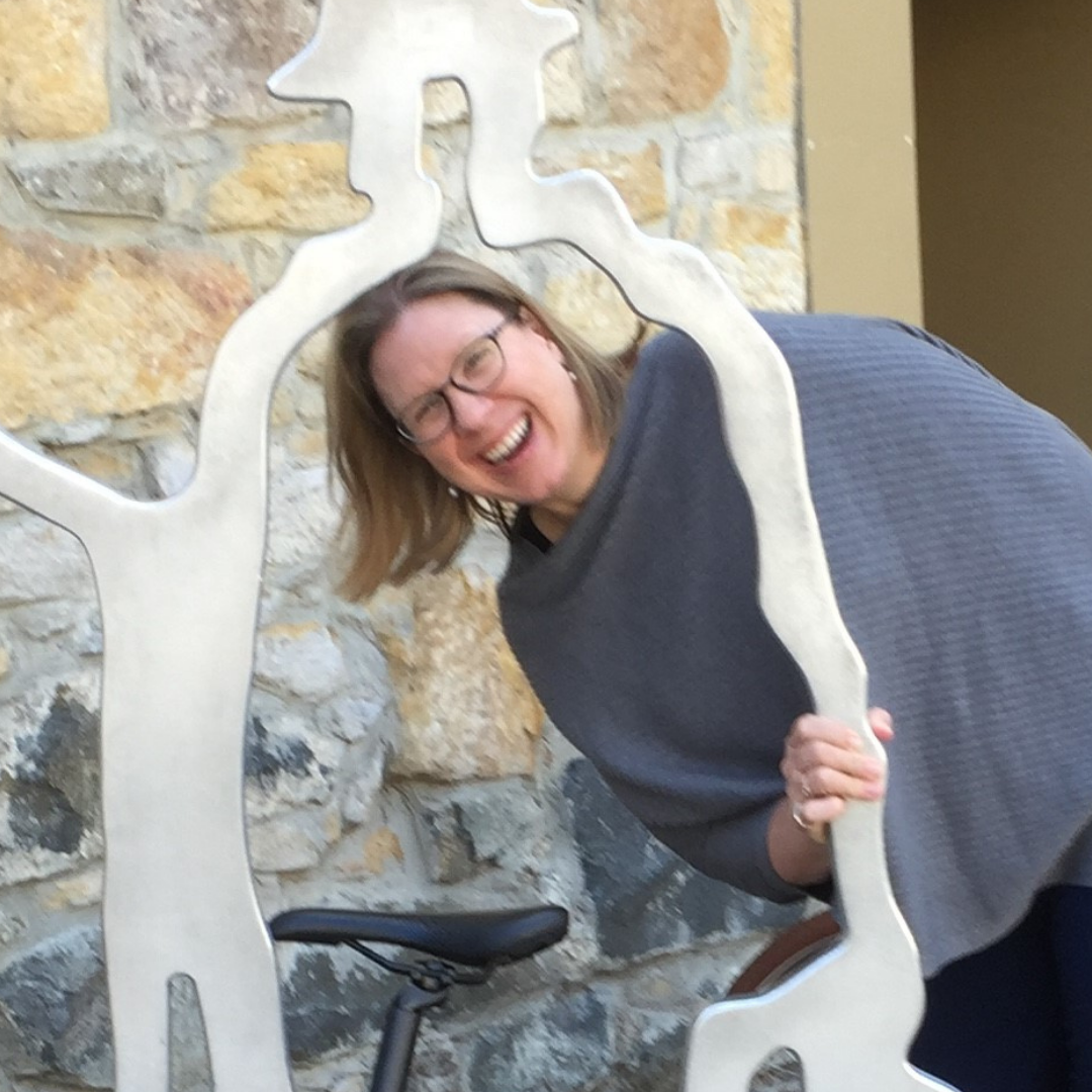 Photo of a smiling white woman with shoulder length blonde hair wearing glasses and a gray sweater posing with her head inside a metal sculpture that is the outline of a person