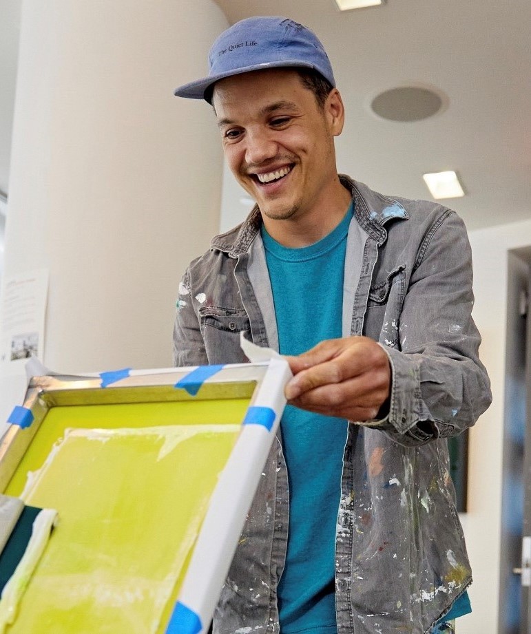Ricardo Roig, professional fine artist and printmaker, smiling wearing a grey flannel with paint splatters on it with a blue t-shirt underneath and a blue cap. He is holding a canvas in the picture and is in an office are with neutral walls.