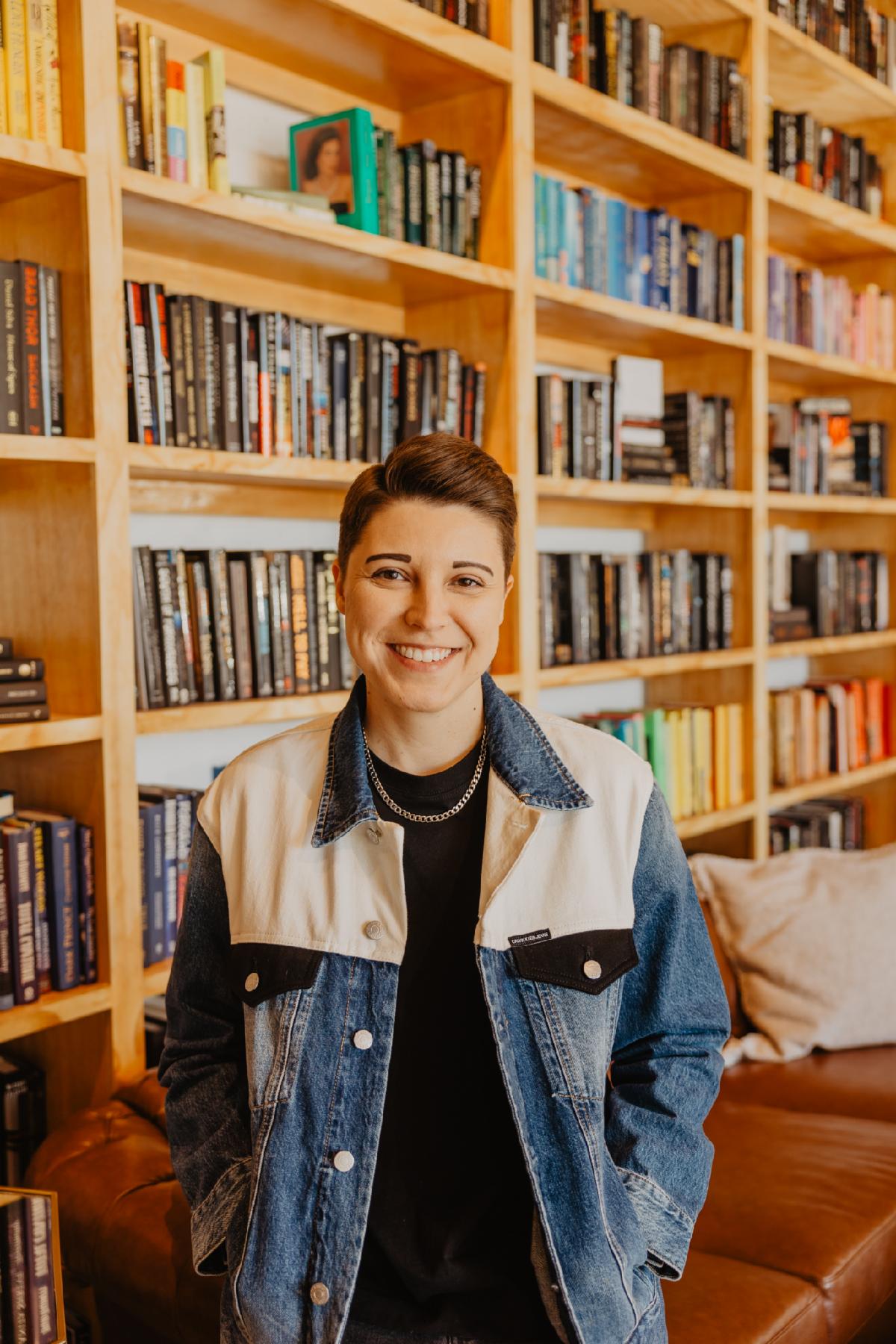 A photograph portrait of smiling person wearing a jacket in a room full of shelves of books.