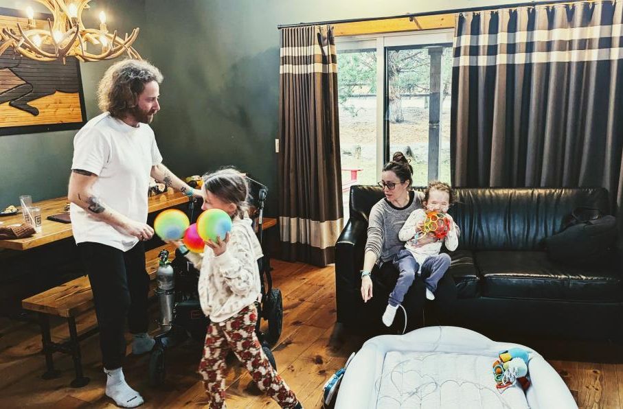 Girl carrying coloured balls walks beside man while woman holds small child on couch