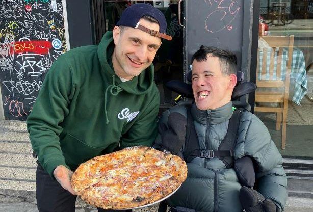 Young man with dark hair in wheelchair grins beside man standing wearing baseball cap and holding a pizza
