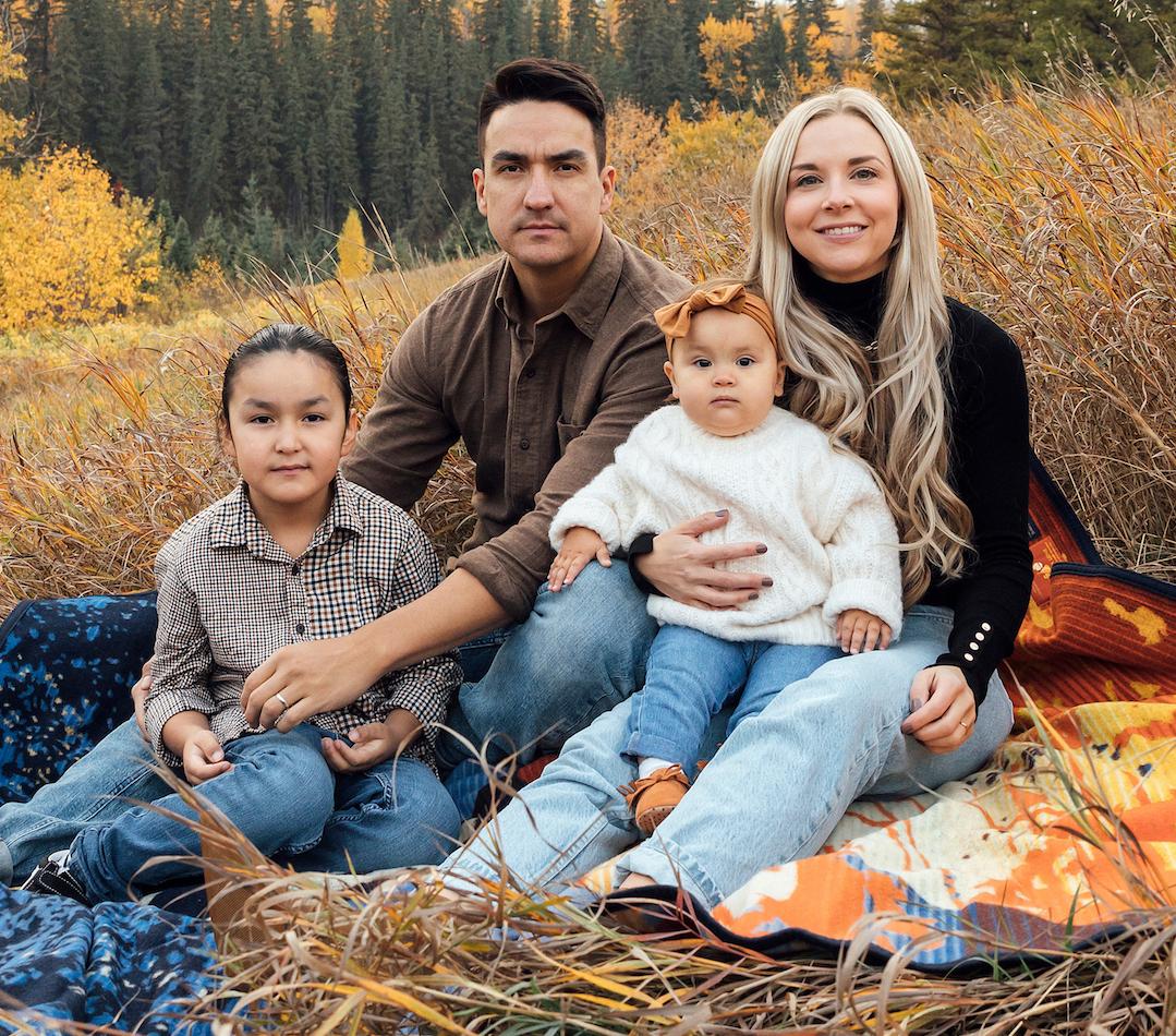 Man with dark hair, woman with long blonde hair and boy and baby girl sit in field