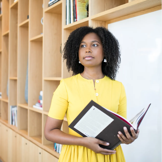 Woman with curly black hair and yellow dress in library holding a book open