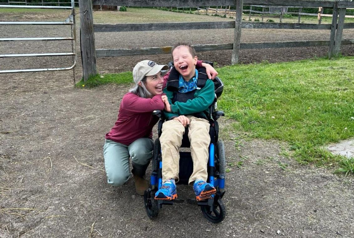 Woman with blonde hair and baseball cap crouches beside boy with green sweatshirt in wheelchair both smiling