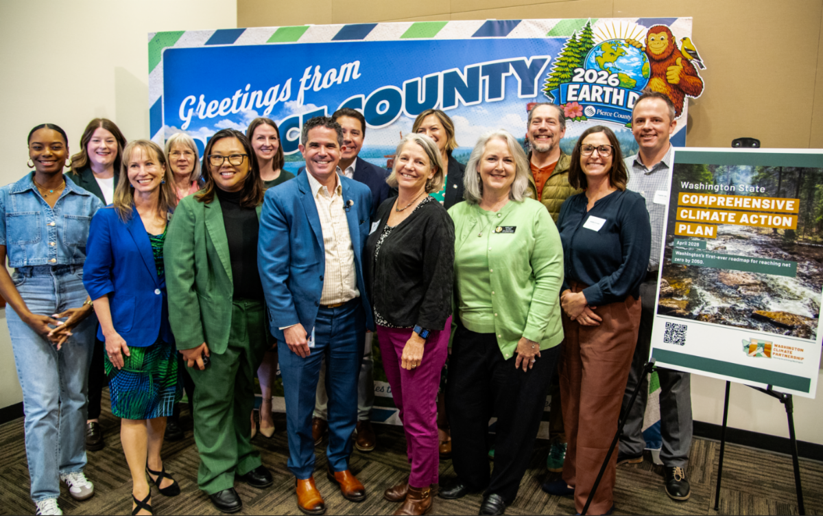 Community leaders from around the state in front of a banner that says "Greetings From Pierce County". Next to a poster that shows the cover of the new Comprehensive Climate Action Plan.