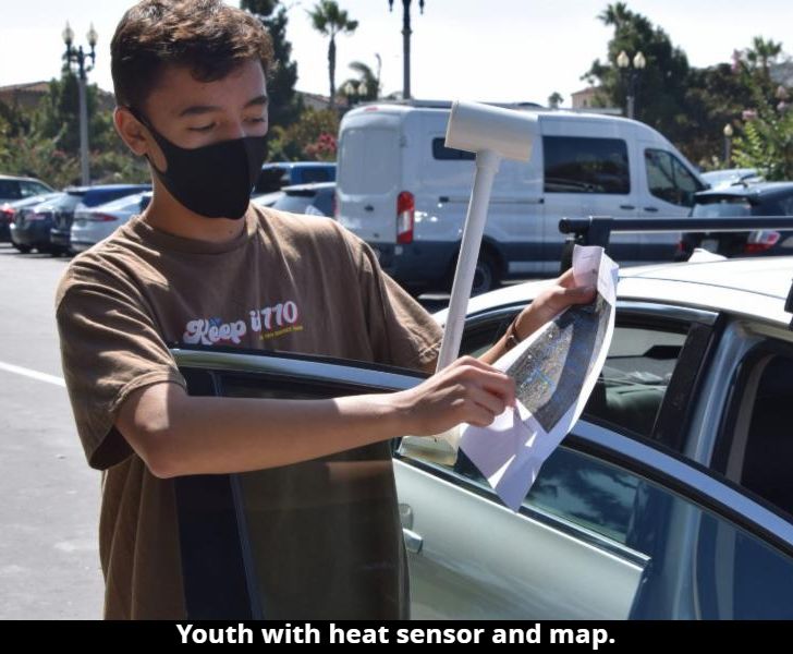 Youth holding a heat sensor and map next to a vehicle with the door open.