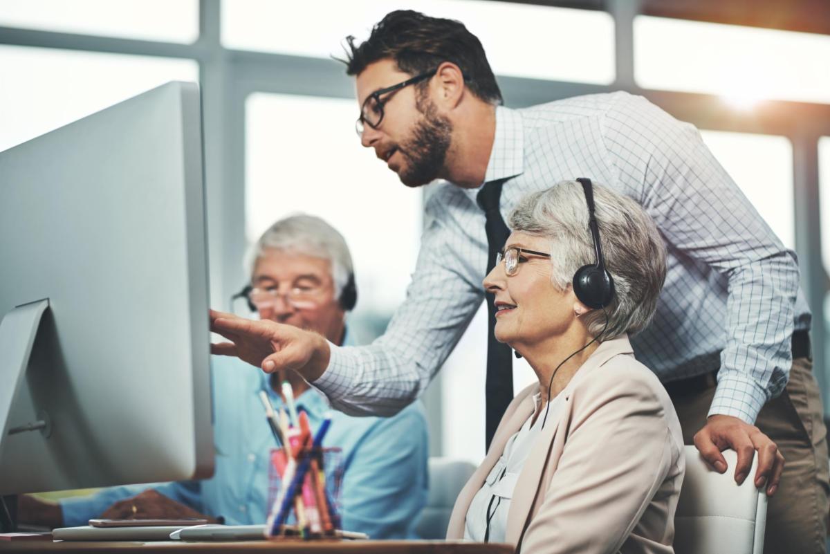 Two older adults seated at a desk, viewing a computer screen and listening through headphones while another adult standing behind them gestures at the screen.  