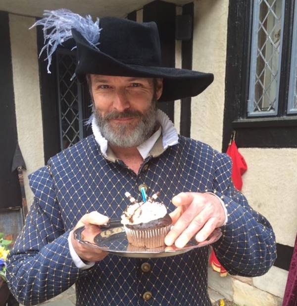 a man wearing a hat and dressed as William Shakespeare holds a cupcake