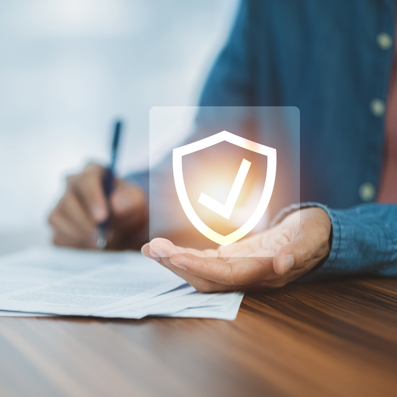 A hand writing with a pen on paper and the other hand holding a superimposed icon of a checkmark in a badge.