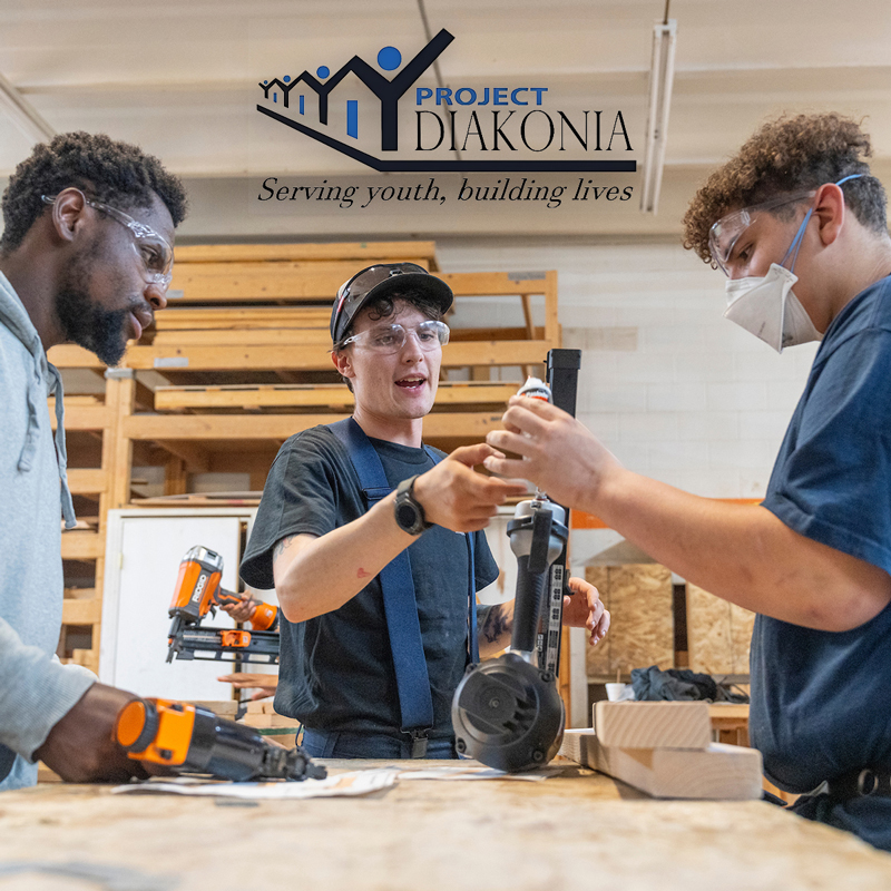 Three students using a woodworking tool inside a Project Diakonia warehouse.