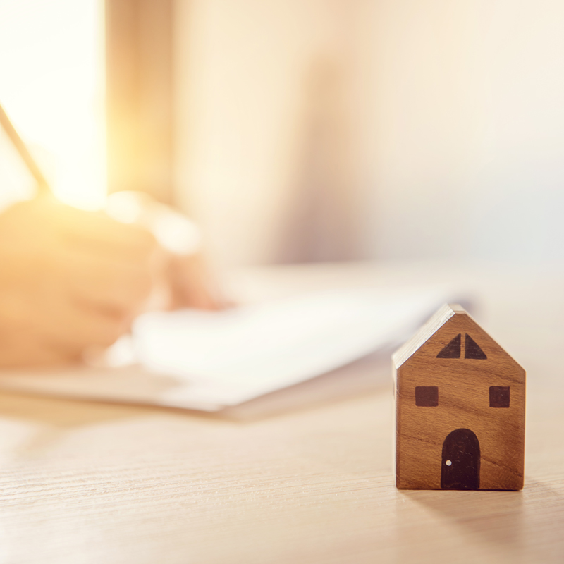 A small wooden house sitting atop a desk which a person writing in a notebook behind it.