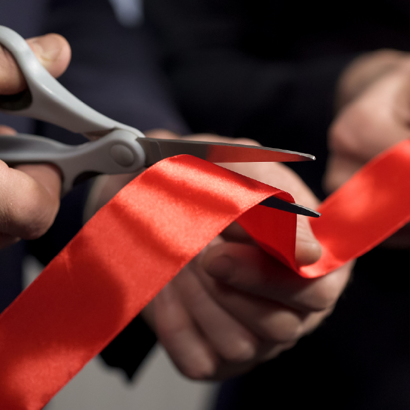 Two sets of hands holding a red ribbon with one hand cutting it with scissors