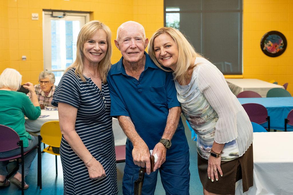 Jay Willey poses for a picture with two women in a room painted with bright yellow walls. The room is set up with multiple banquet tables. 