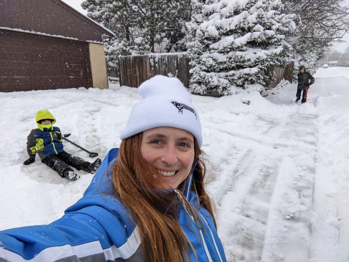 A close-up selfie of a smiling woman wearing a white beanie and a blue winter jacket on a snowy driveway. In the background, a young child in a bright yellow and blue snowsuit sits in the snow near a shovel, while another person stands further up the snow-covered street near pine trees