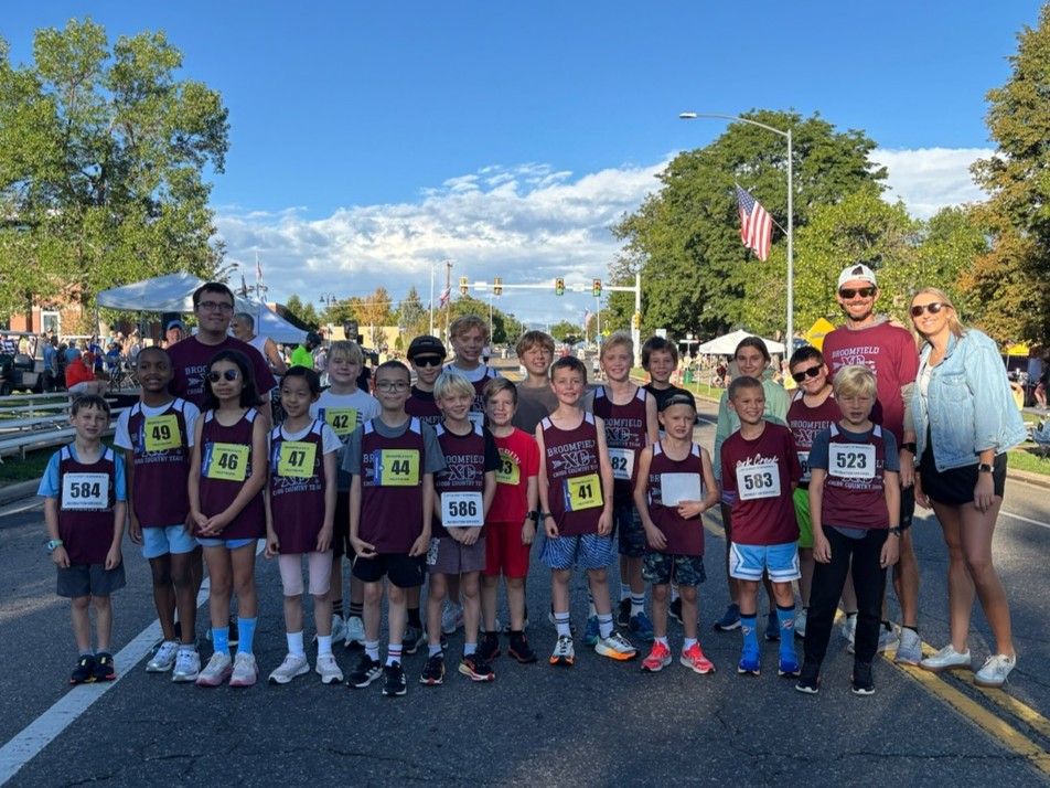 A large group of young CARA Cross Country athletes posing outdoors on a paved street during Broomfield Days. The kids wear matching maroon and white race jerseys with bib numbers attached. Adult coaches stand alongside the group under a clear blue sky.