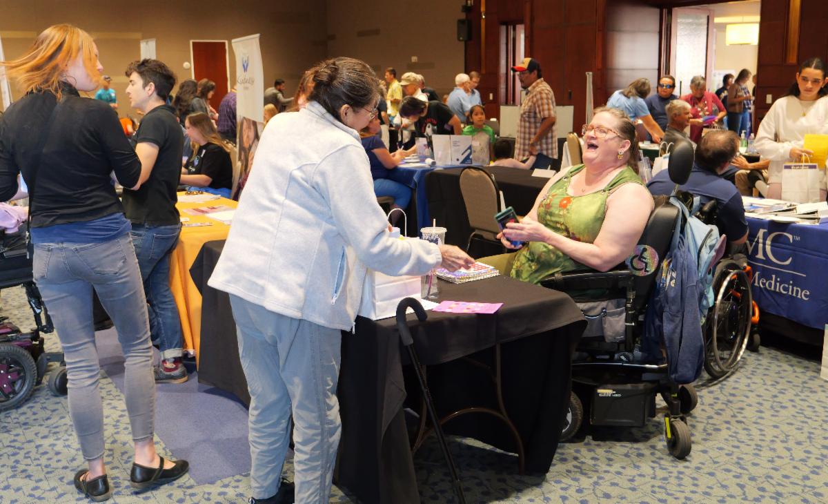 group of people gathered around exhibitor tables at the resource fair. 2 people in wheelchairs are in the image.