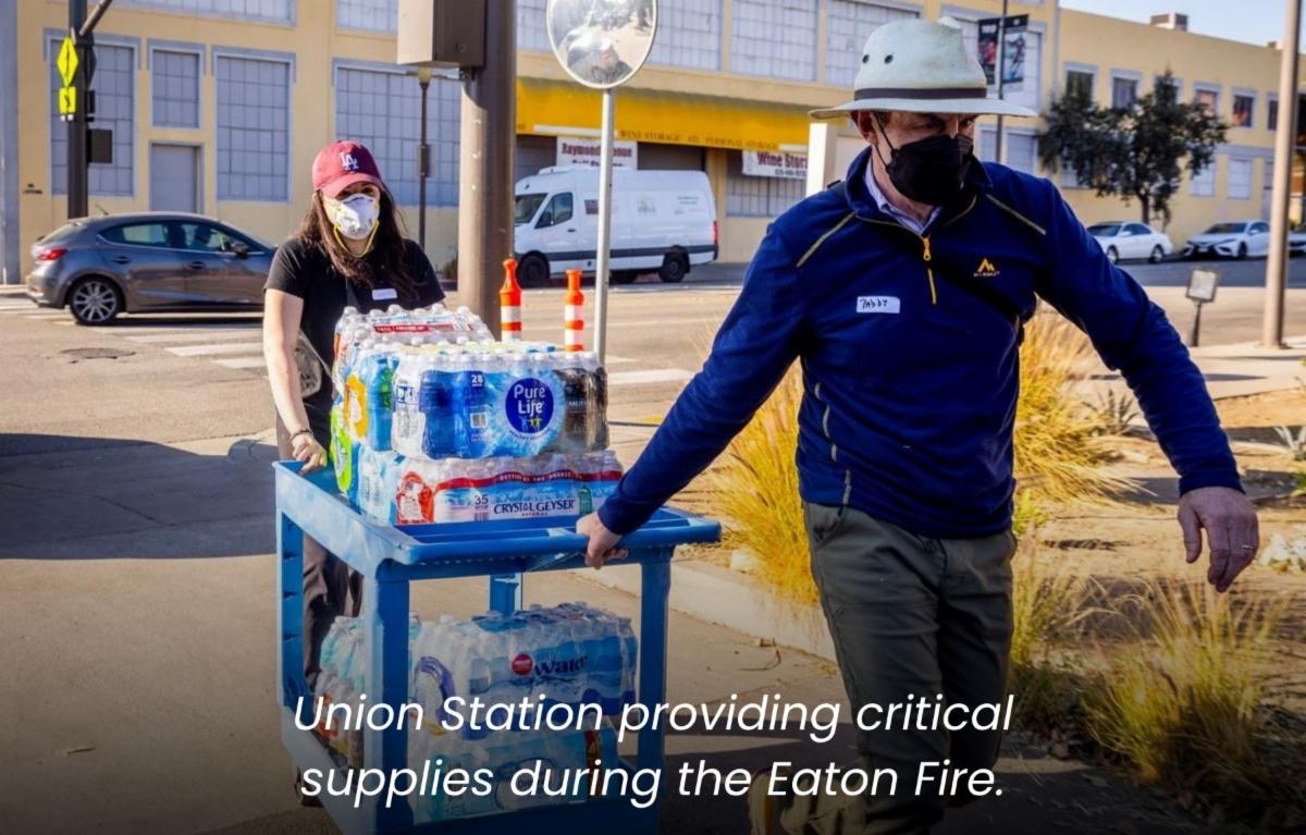 a woman in a mask pushes a cart full of water while a man in a white hardhat pulls the cart