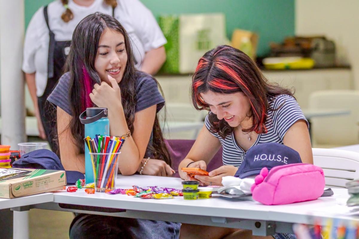 two young women seated at a table covered in craft supplies, while one looks at her phone and the other looks over her shoulder 