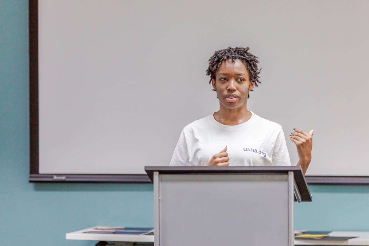 a woman in a white t-shirt speaking at a podium in front of a projector screen