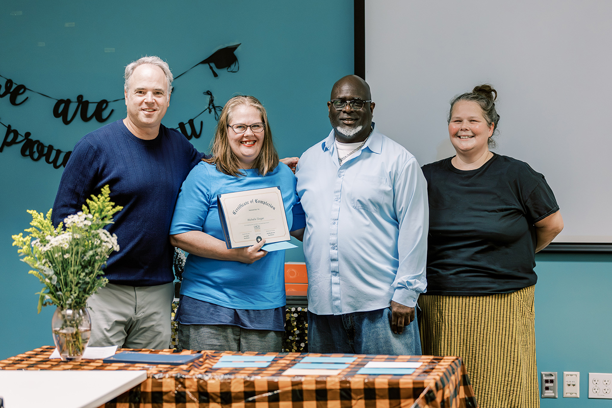 a group of two women and two men standing in front of a blue wall, while one woman holds up a certificate