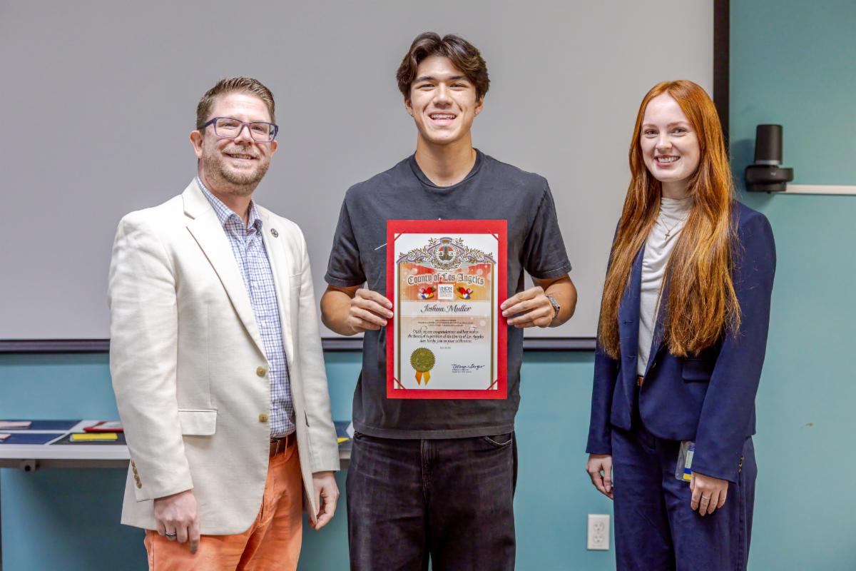 a young man holds a certificate while standing between a young woman with red hair and a man in a white jacket 