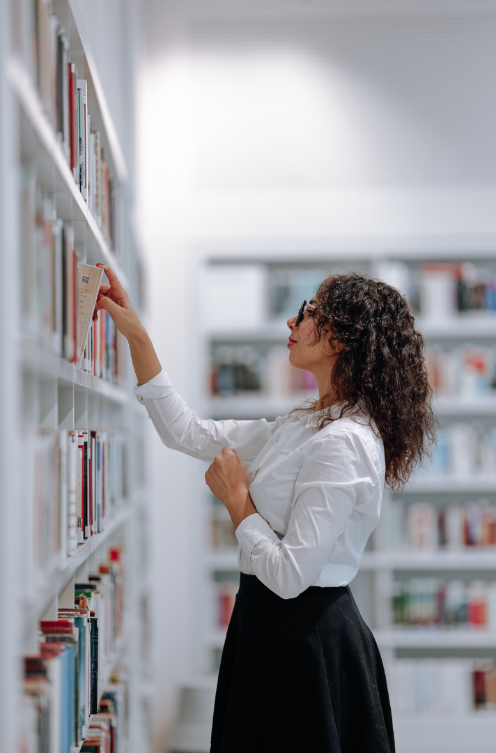 Photo of a woman in a white blouse and black skirt looking at a shelf of library books and choosing a book