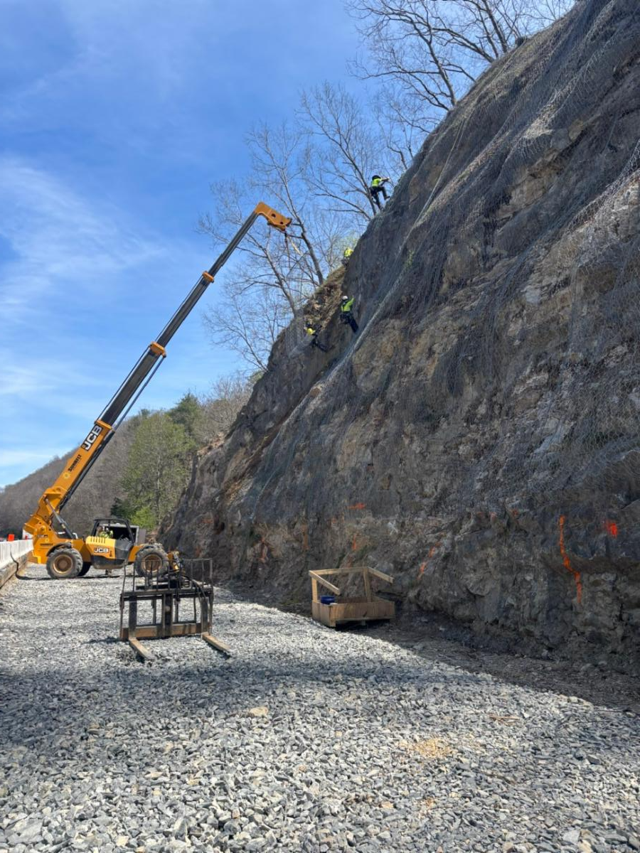 Crane beside rock wall with people in PPE rappels down rock slope covered with a rock drape to prevent rocks from entering road