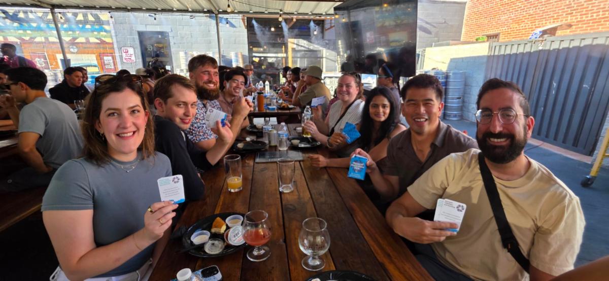 Resident and fellow physicians seated around a large wooden table at an outdoor networking mixer, holding Dynamic Discussions Deck cards as part of a facilitated conversation activity. Food, beverages, and discussion cards are visible on the table, while additional attendees participate in conversations at nearby tables in the covered patio setting.