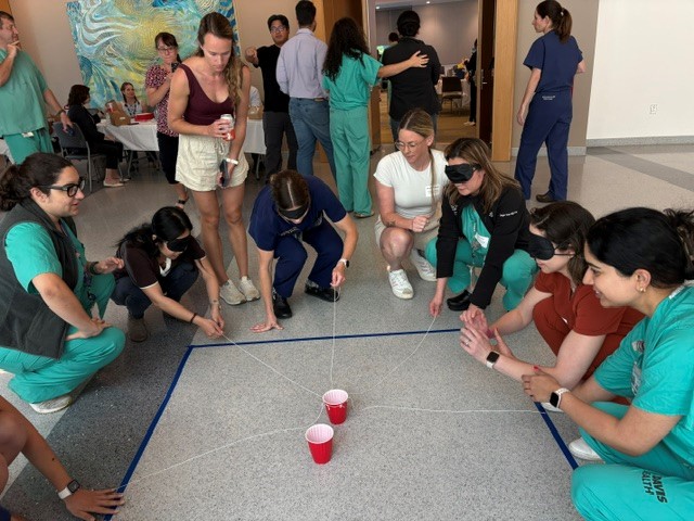 semi-circle of eight people kneeling, holding strings, blindfolded, with cups in the center.