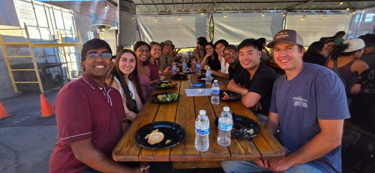 Residents and fellow physicians seated around a long wooden picnic table at an outdoor networking mixer. Participants are engaged in group conversation while sharing a meal, with water bottles, plates, and discussion materials on the table. The gathering takes place under a covered patio with string lights, and additional attendees are seated at nearby tables in the background.