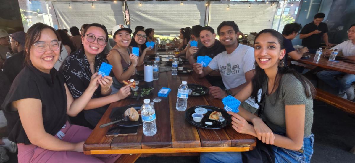 Resident and fellow physicians seated around a wooden picnic table at an outdoor networking mixer, holding Dynamic Discussions Deck cards while participating in a guided conversation activity. Food and water bottles are on the table, and additional attendees are gathered at nearby tables under a covered patio with string lights.