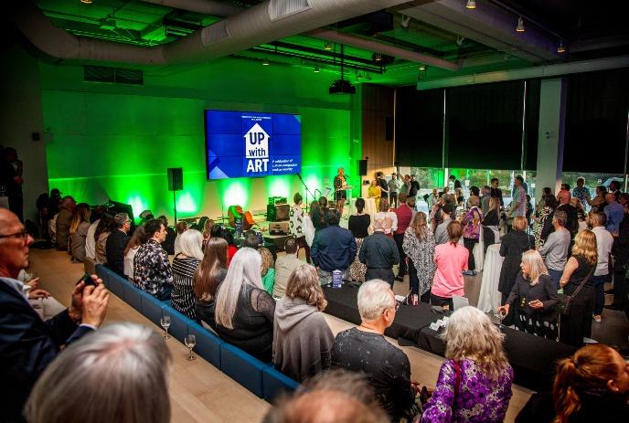 From tourismlondon.ca: Crowd gathered in a modern auditorium at Museum London with green lighting for an event titled 'UP with ART' displayed on a large screen.