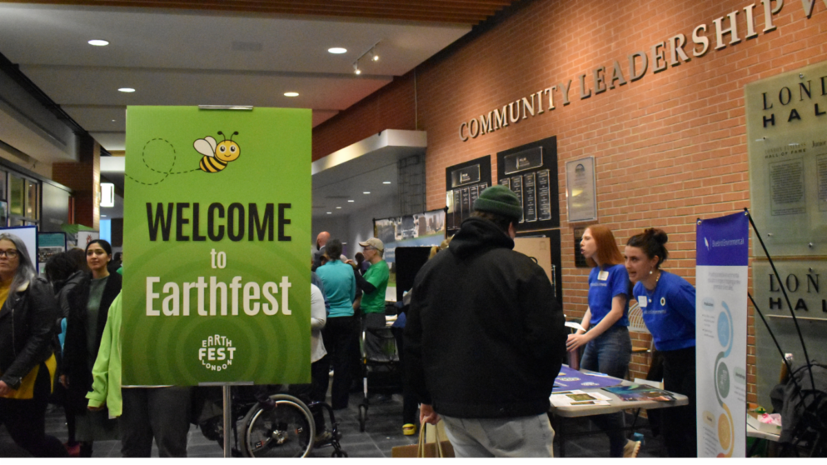 From earthfestlondon.ca: Green sign with a bee graphic welcoming attendees to London's Earthfest at a busy indoor event with people interacting at tables.