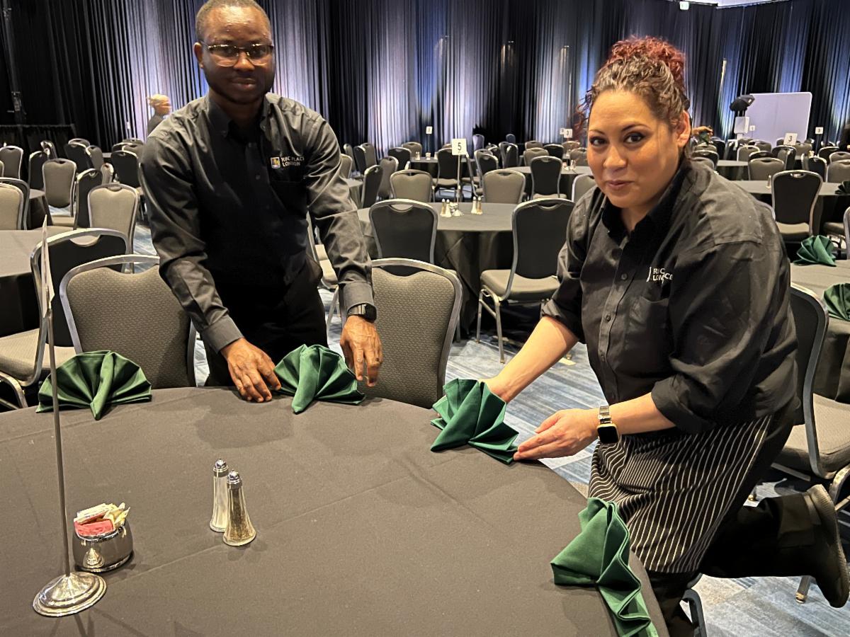 A seasoned event services associates demonstrates proper folding technique to a new team member during setup of the Ivey Formal event.